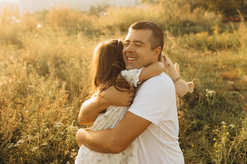 `Smiling father hugging his young daughter in a grassy field during golden hour backlight.`